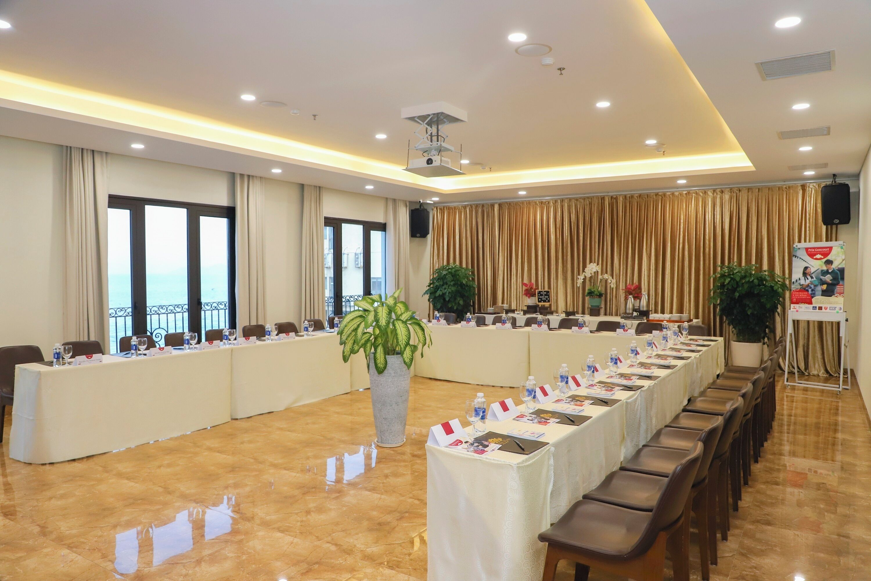 A conference room with a long table, chairs, a large potted plant, and a speaker on the wall.