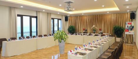 A conference room with a long table, chairs, a large potted plant, and a speaker on the wall.