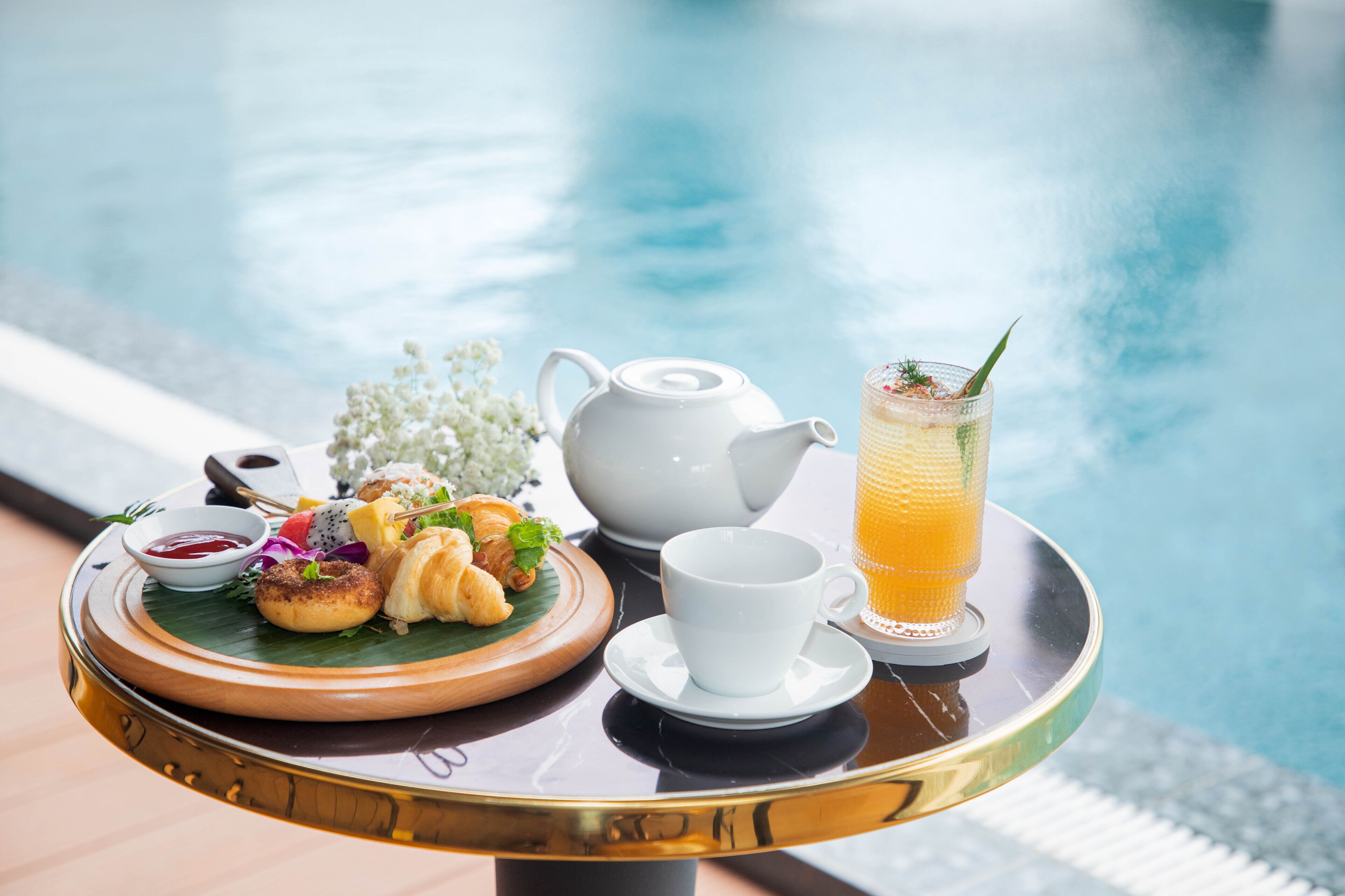 A tray with croissants, a teapot, a cup, and a glass of orange juice by a pool.