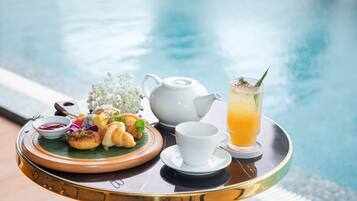 A tray with croissants, a teapot, a cup, and a glass of orange juice by a pool.