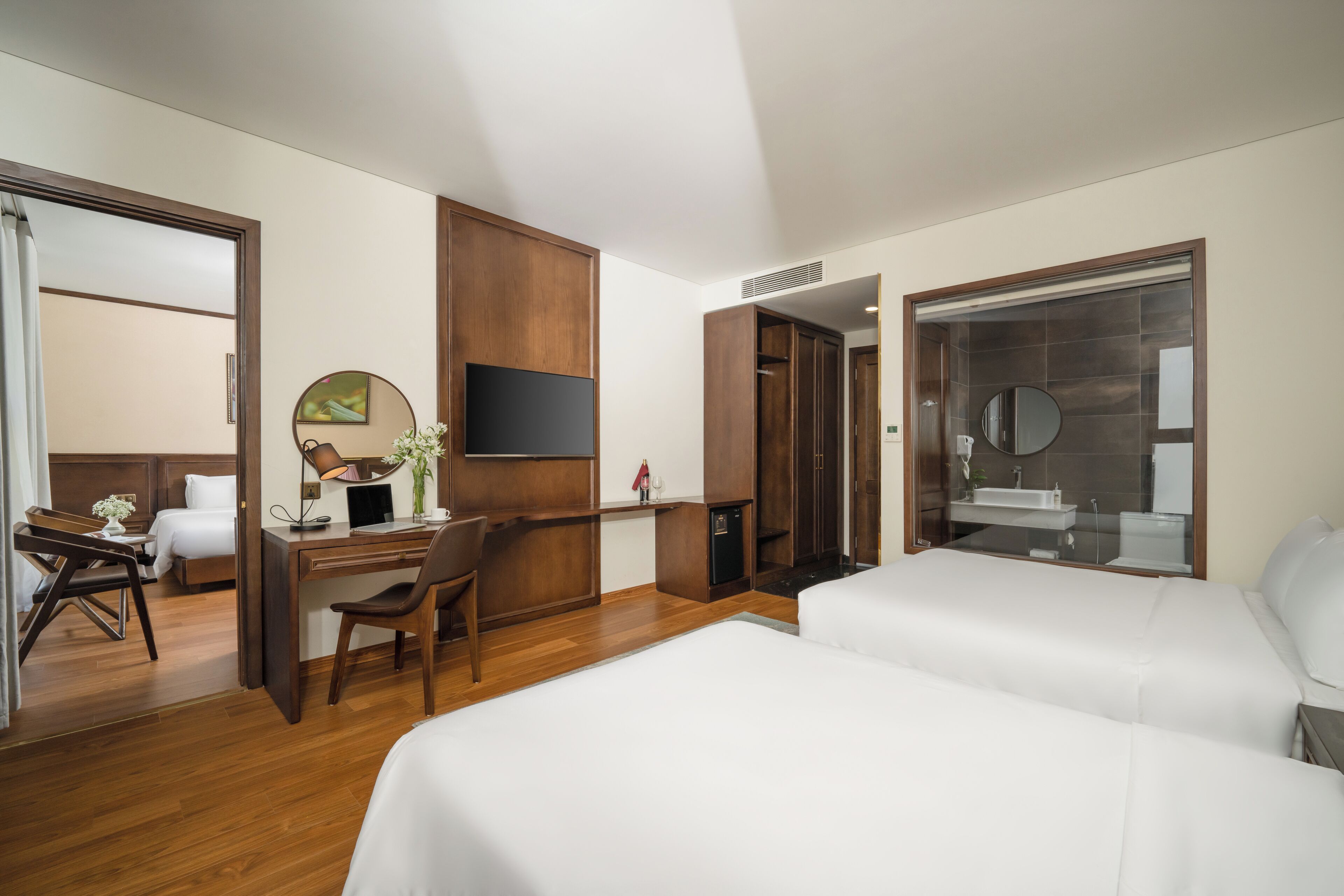 A hotel room with two beds, a desk with a chair, a TV, and a bathroom with a mirror and sink.