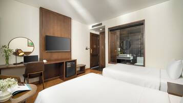 Quarto de hotel com cama, escrivaninha com computador, cadeira, mini-geladeira, televisão e banheiro visível através de uma porta de vidro.