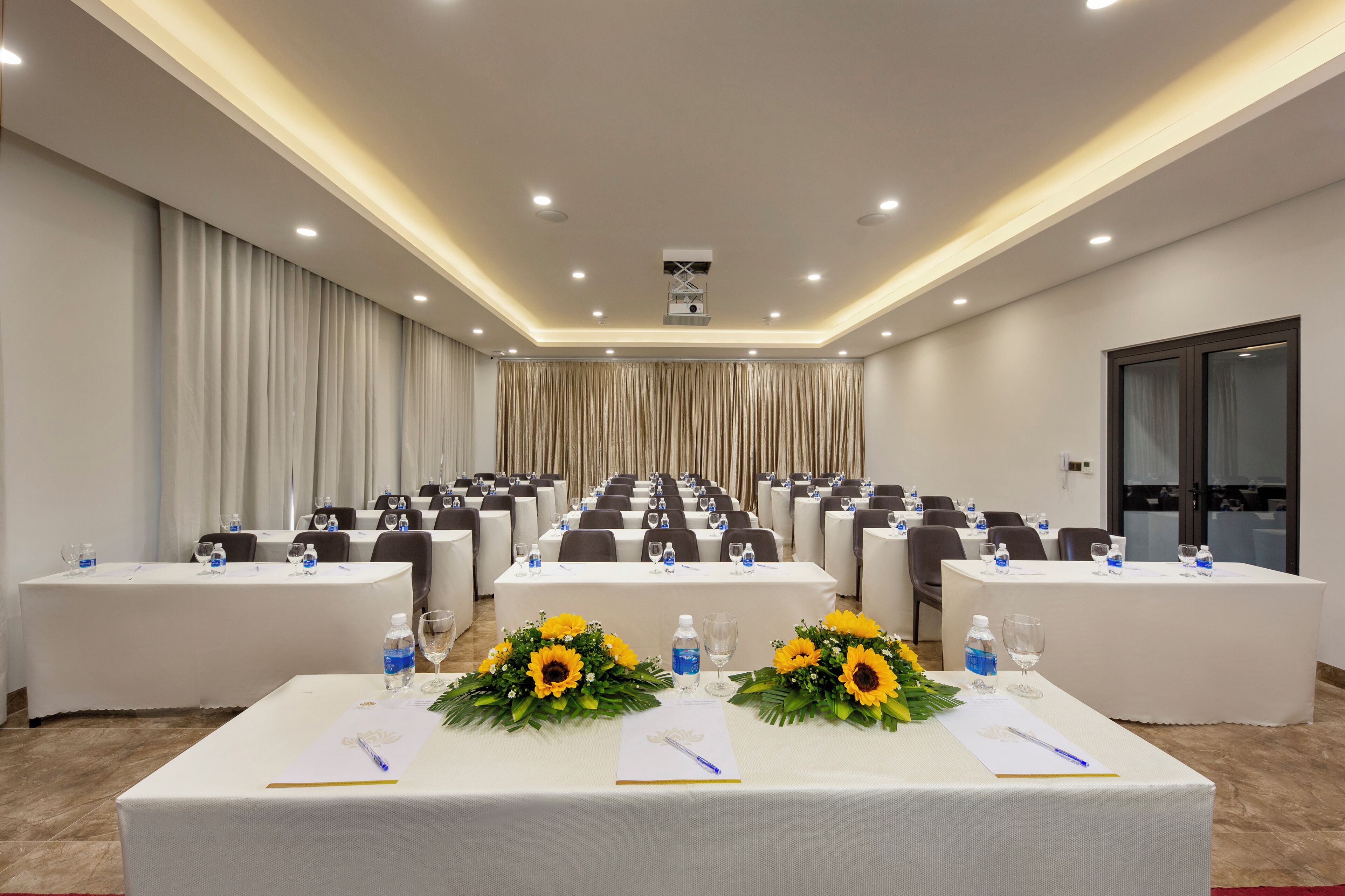 A conference room with rows of tables, each with water bottles, glasses, and sunflowers.