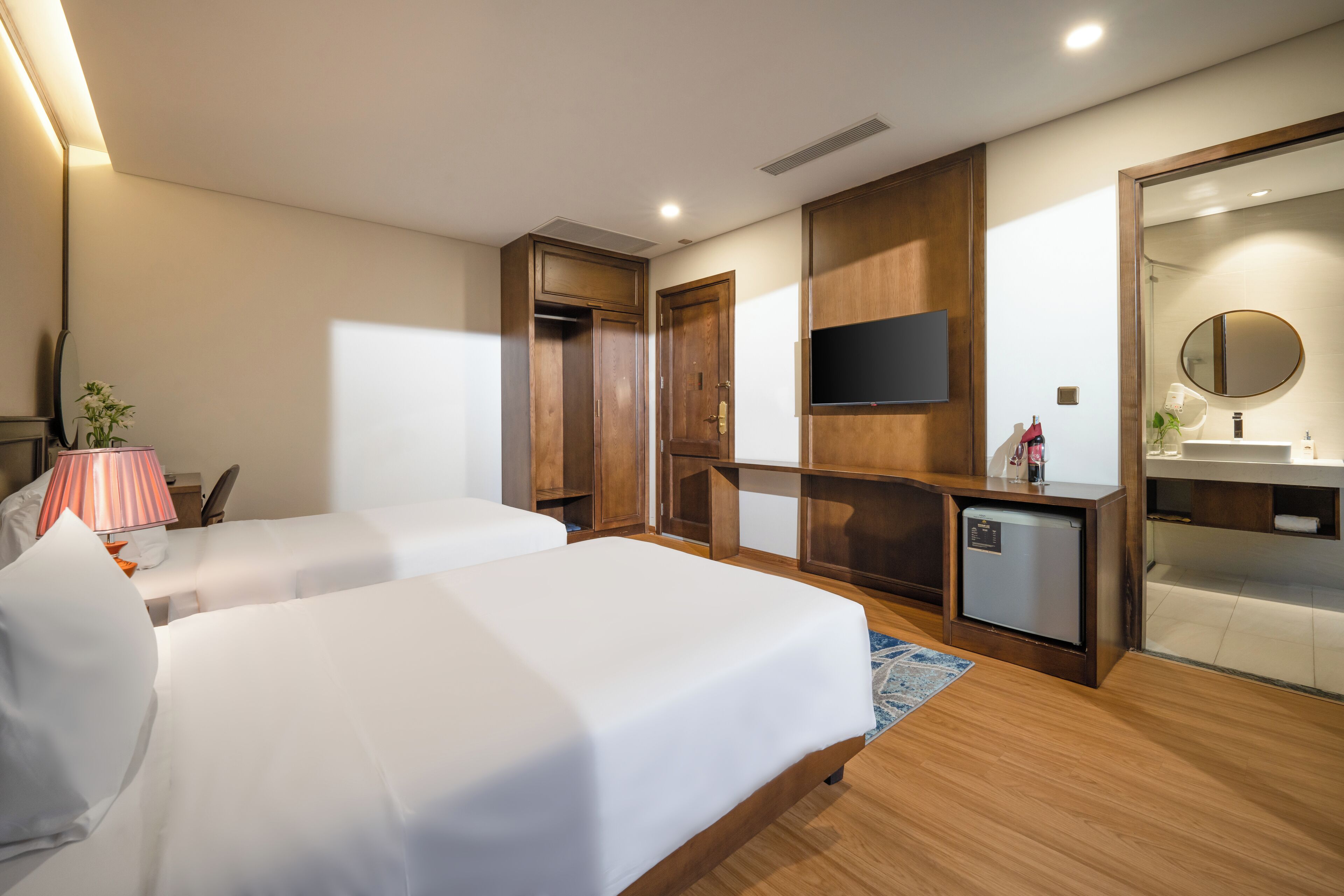 A hotel room with two beds, a TV, a small kitchenette, and a bathroom with a mirror.