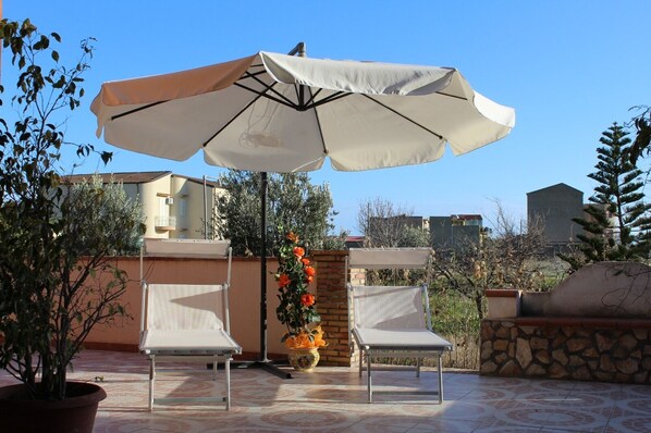 An outdoor patio with two white chairs, a large umbrella, and a potted plant with orange flowers.