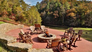 Un pozo de fuego de piedra circular rodeado de sillas Adirondack de madera, ubicado en un patio rojo con un muro de piedra y vistas a un campo de golf y un bosque.