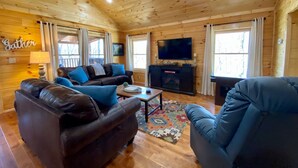 Interior acogedor de una cabaña de troncos con chimenea, un televisor y un cartel que dice 'Gather'.