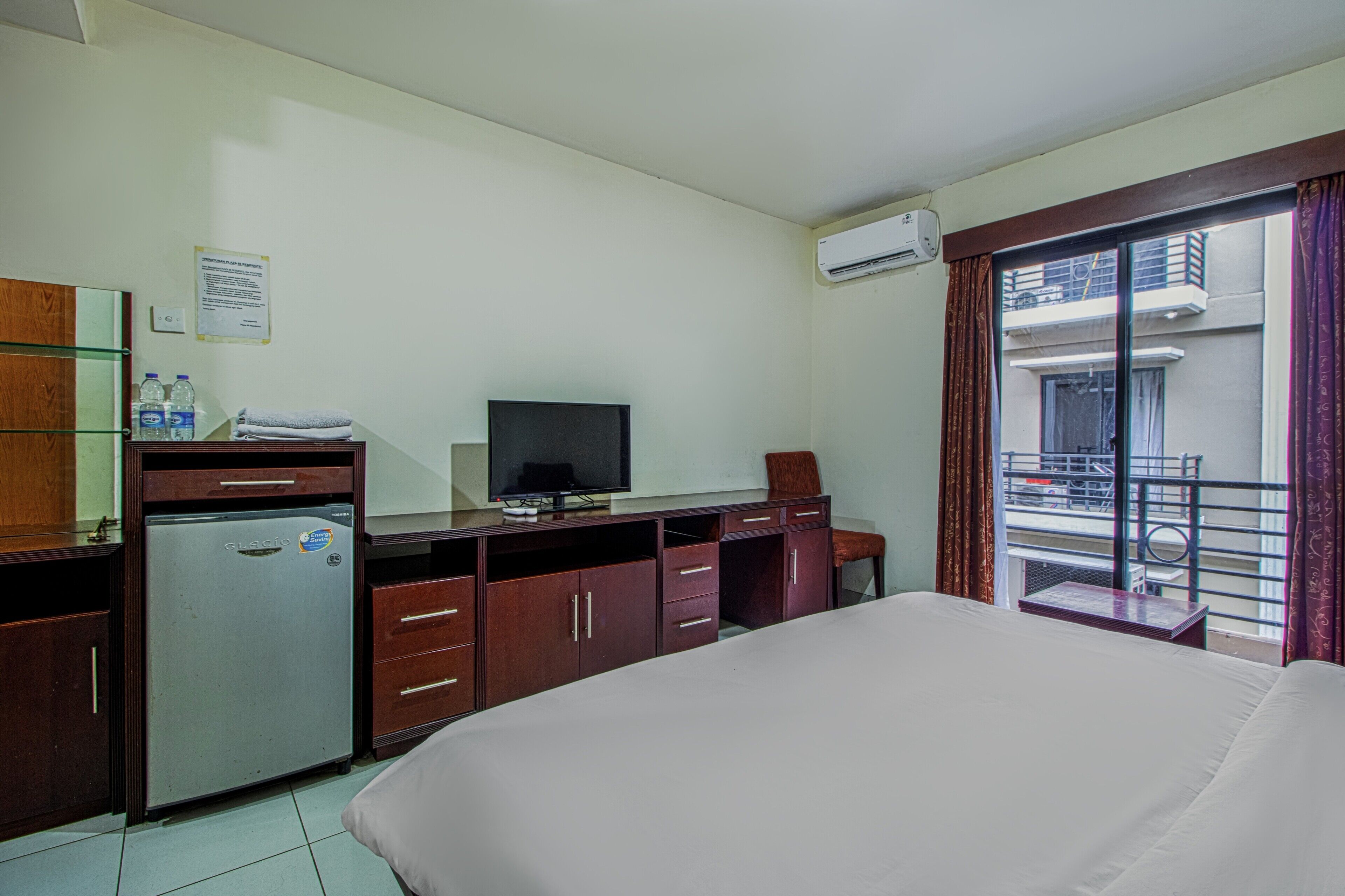 A hotel room with a bed, a desk with a television, a refrigerator, and a window with a view of the outside.