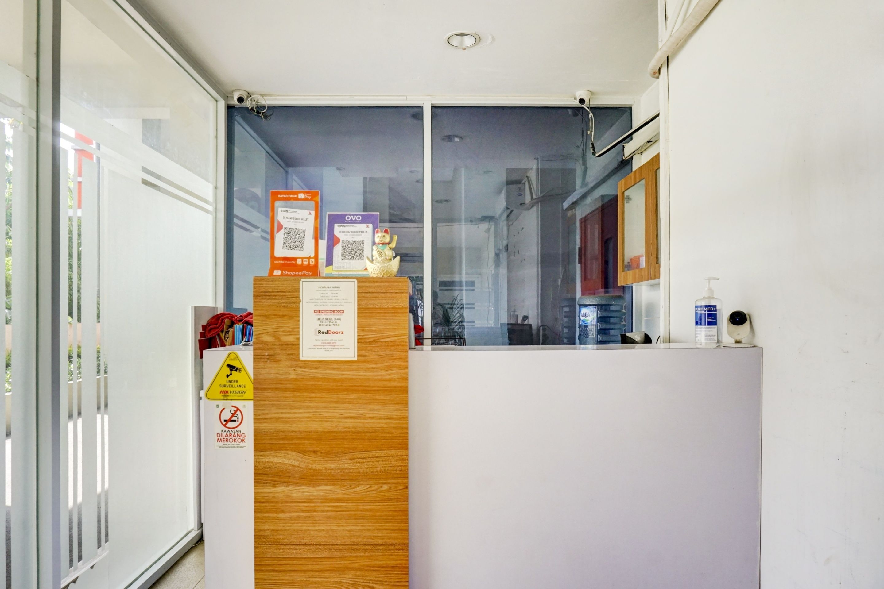 Camera d'albergo con un divisorio in legno, vista attraverso una porta in vetro e diversi cartelli, tra cui un segnale di divieto di fumo e un avviso di videosorveglianza.