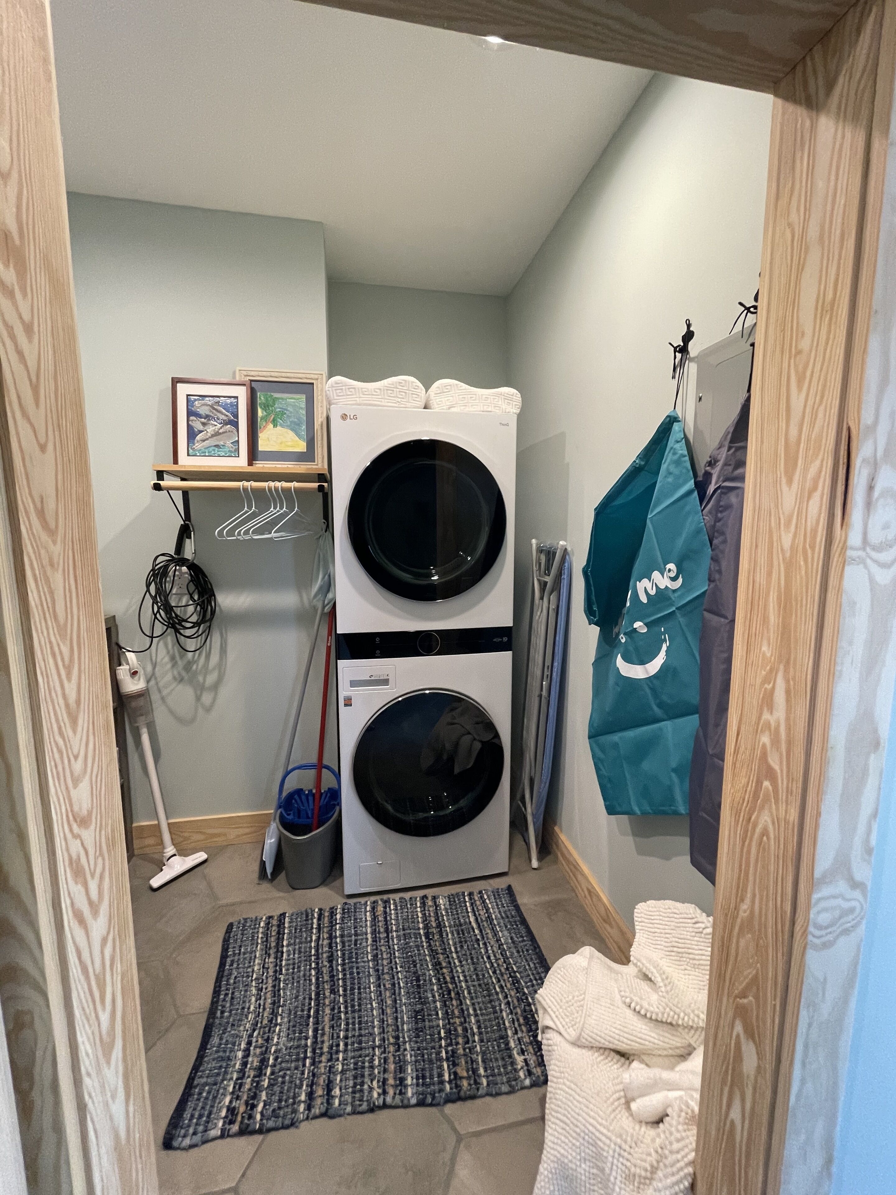 A laundry room with two stacked washing machines, a drying rack, a vacuum cleaner, and a blue mat on the floor.