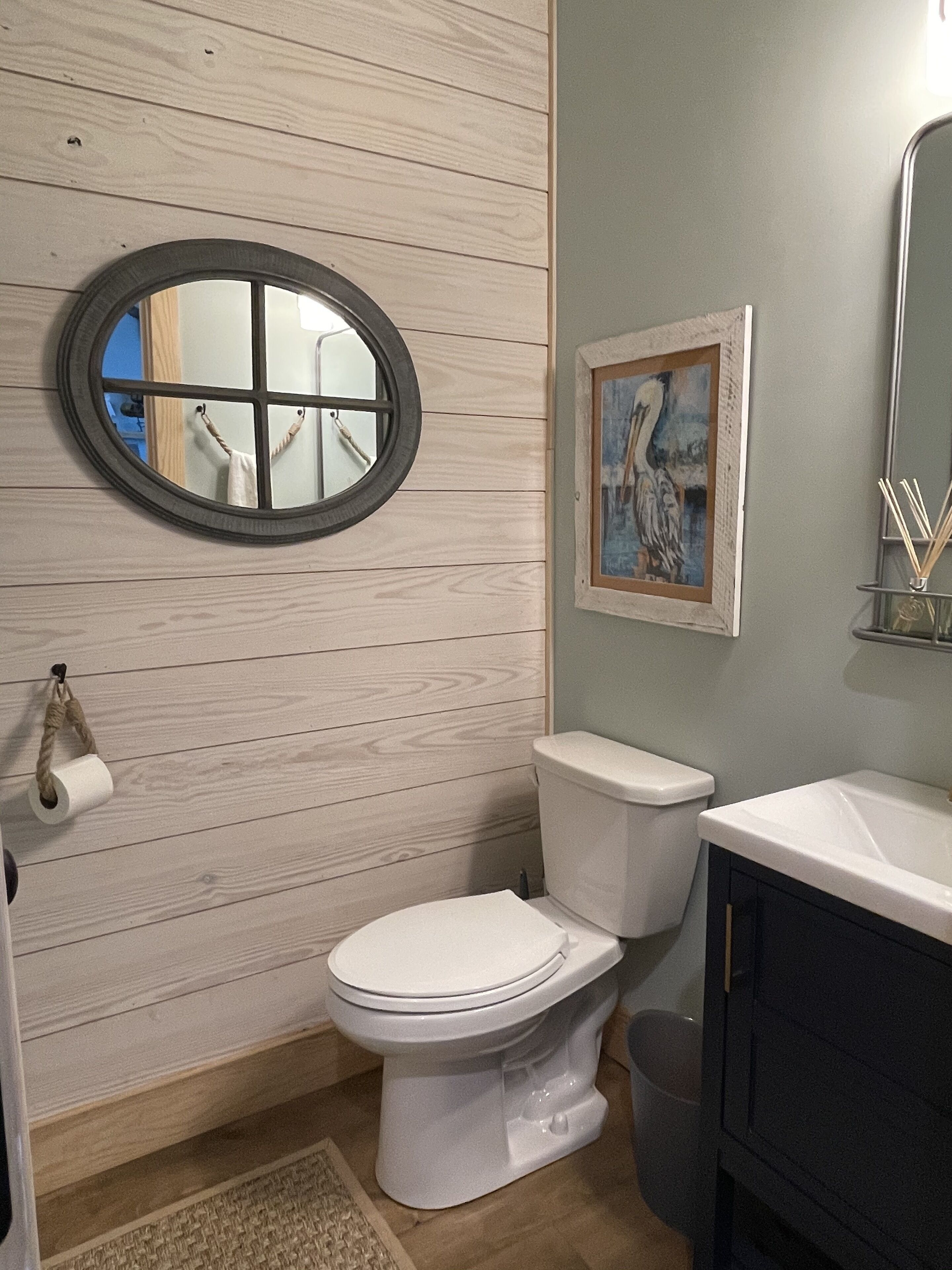 Une salle de bain avec un mur en bois, un miroir rond, des toilettes, un lavabo et un tableau encadré.