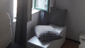 Ein Zimmer mit Fenster, einem weißen Stuhl, einem gemusterten Kissen und einem schwarz-weiß gemusterten Sitzkissen.