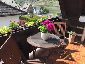 Une terrasse avec une table ronde et deux chaises en bois, entourée de plantes en pot et offrant une vue sur une rivière et des maisons au loin.