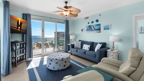 A living room with a sofa, ottoman, and chairs, a ceiling fan, and a view of the ocean through a sliding glass door.