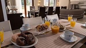 Une table dressée pour le petit-déjeuner, avec des viennoiseries, du jus d’orange et des tasses de café, dans une pièce bien éclairée qui offre une vue sur l’extérieur.
