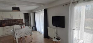 Zona pranzo con tavolo e sedie, cucina con pensili, televisore fissato al muro e finestra con tende.