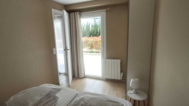 Quarto com cama, radiador e vista para árvores por uma porta aberta.