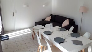 Uma área de refeições com uma mesa branca posta para quatro pessoas, um sofá preto com almofadas e um abajur de chão.