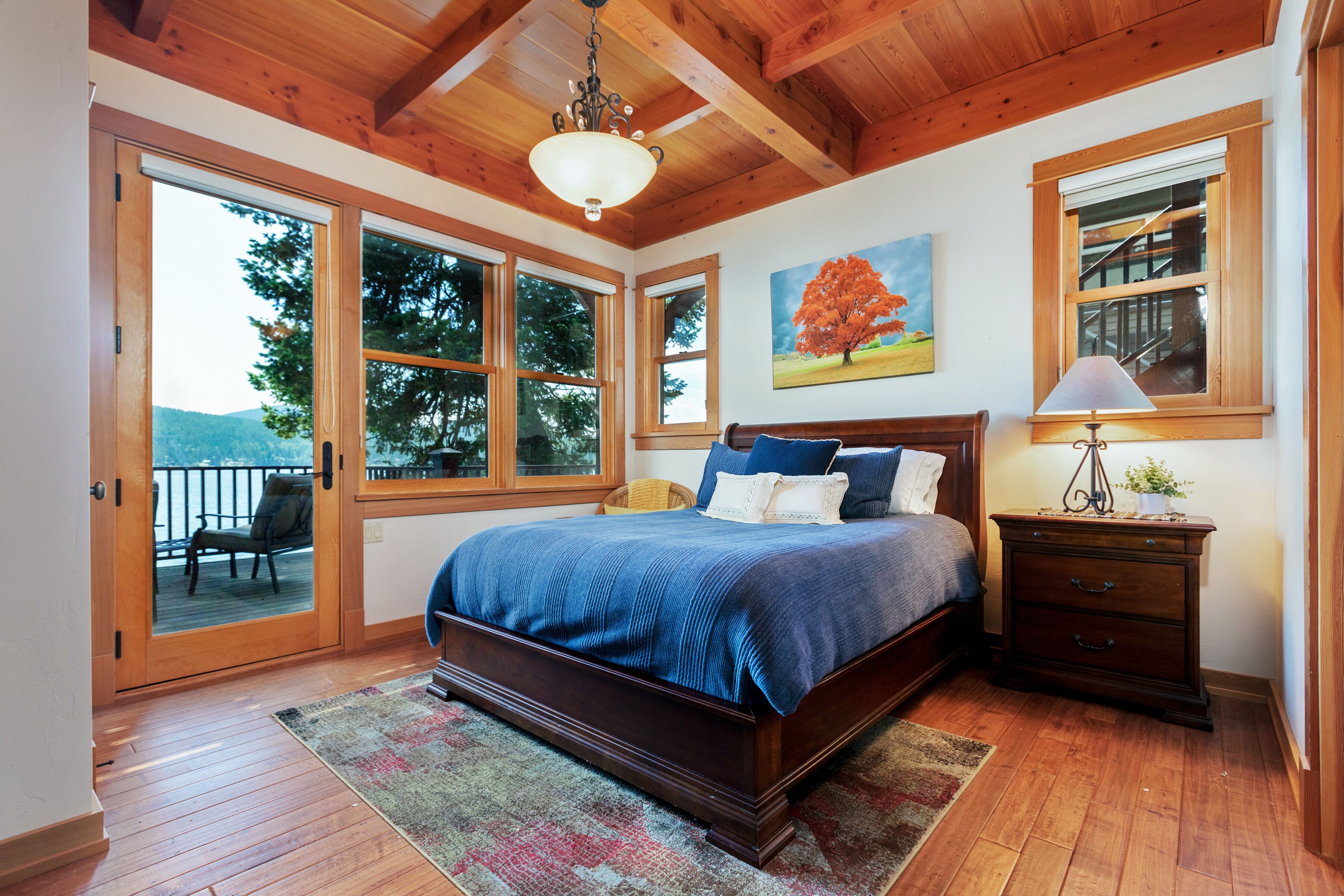 A bedroom with a large bed, wooden flooring, a chandelier, a painting, and a window with a view of trees.