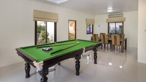 Una mesa de billar en una habitación con zona de comedor, ventanas amplias y un equipo de aire acondicionado.