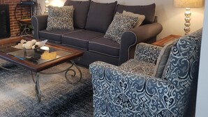 Ein Wohnzimmer mit einem dunkelbraunen Sofa, gemusterten Sessel, einem Couchtisch mit Dekoration, Wandleuchten und einem gerahmten Bild an der Wand.