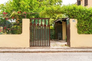 Une entrée fermée par un portail en métal, un chemin en béton et un bâtiment sur lequel est apposée une enseigne indiquant l’adresse.