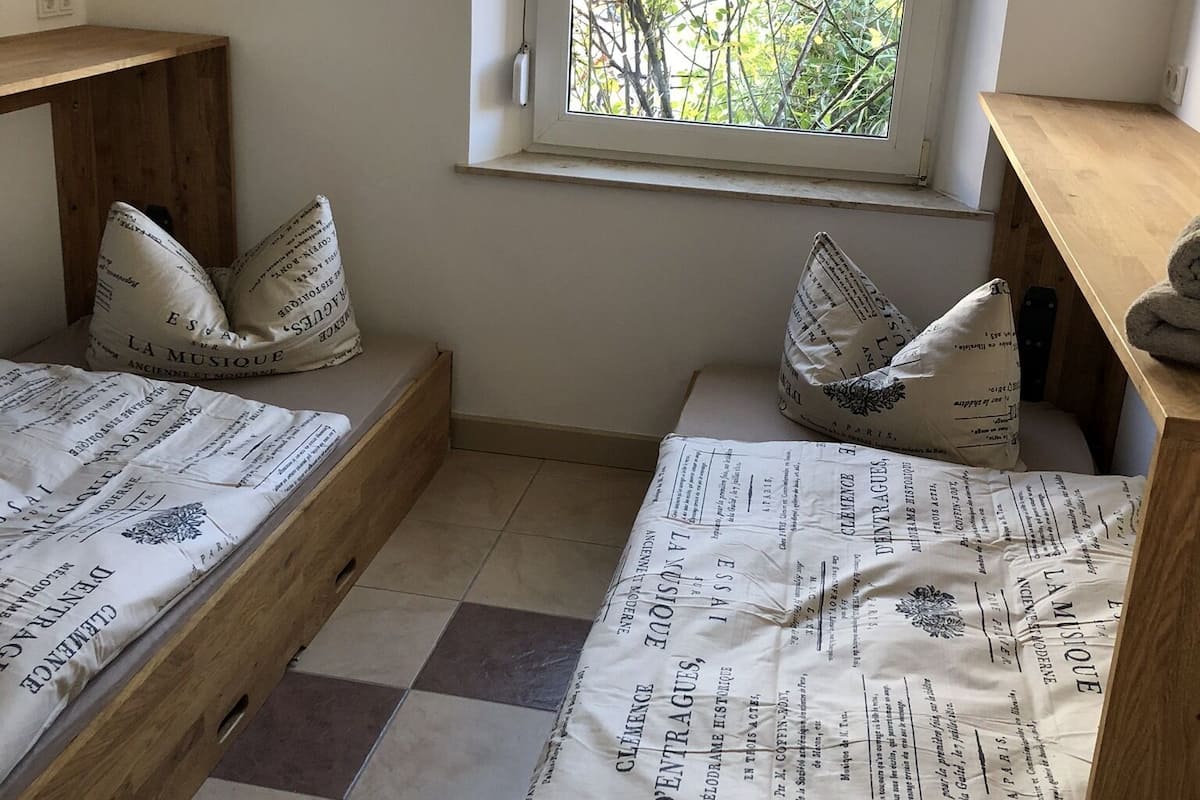 A small room with two wooden beds, each with patterned bedding and pillows. There is a window with a view of trees outside.