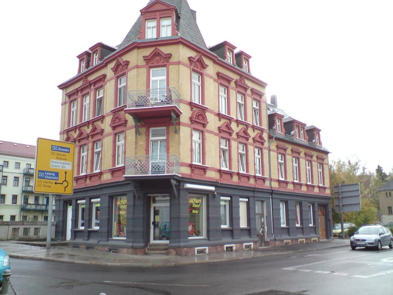 Ein mehrstöckiges Gebäude mit Hauseingang an einer Ecke, einem Geschäft im Erdgeschoss und einem Straßenschild, das auf nahegelegene Ziele hinweist.