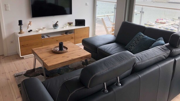 Una sala de estar moderna con un televisor grande, un mueble bajo de madera, un sofá de cuero negro y vista al mar.