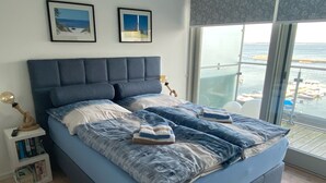 Un dormitorio con una cama grande, una mesita de noche con libros, un ventanal con vista al mar y dos cuadros enmarcados en la pared.