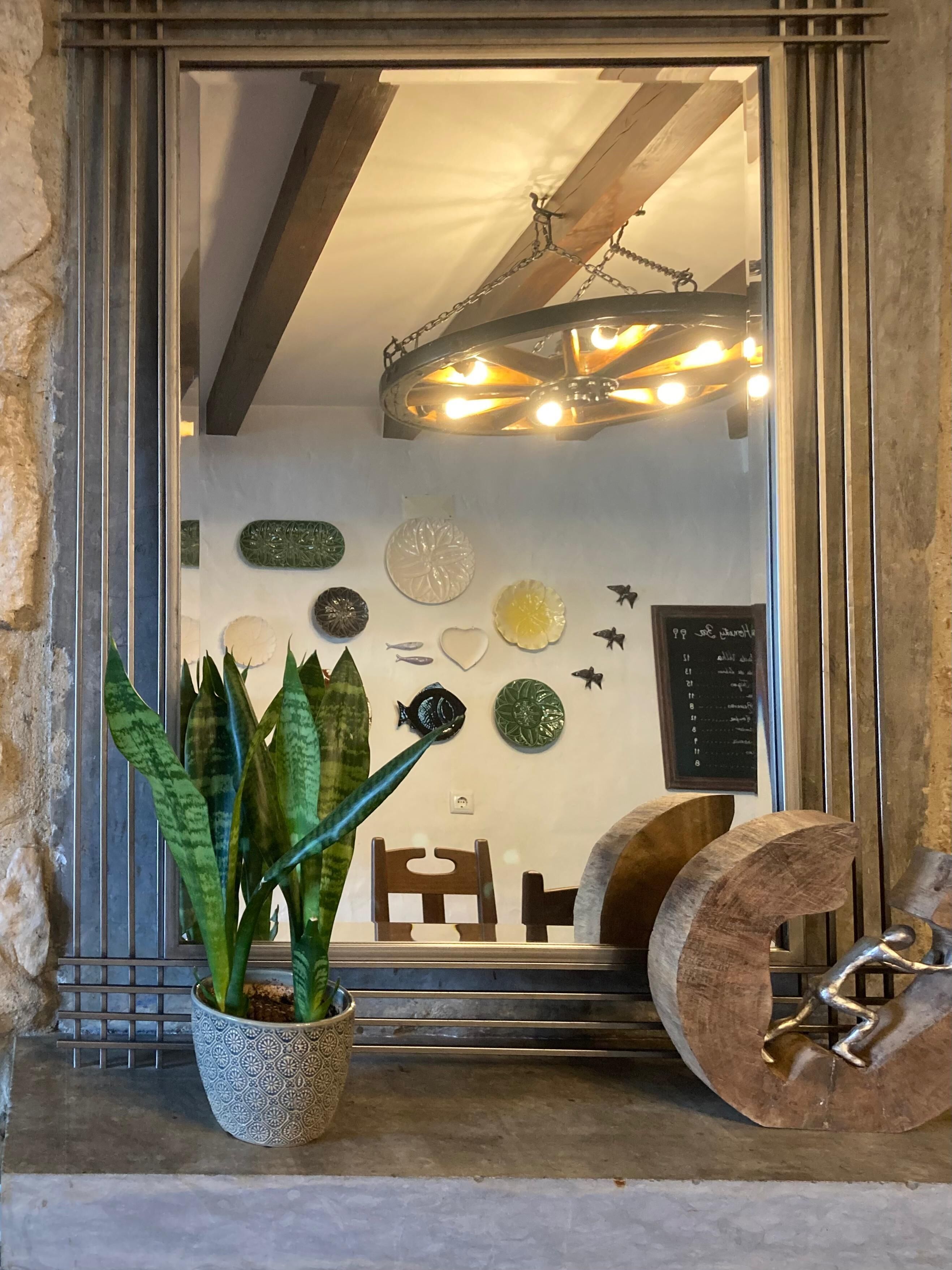 Un miroir reflétant une pièce avec un luminaire au plafond, une plante en pot et des œuvres d’art décoratives accrochées au mur.