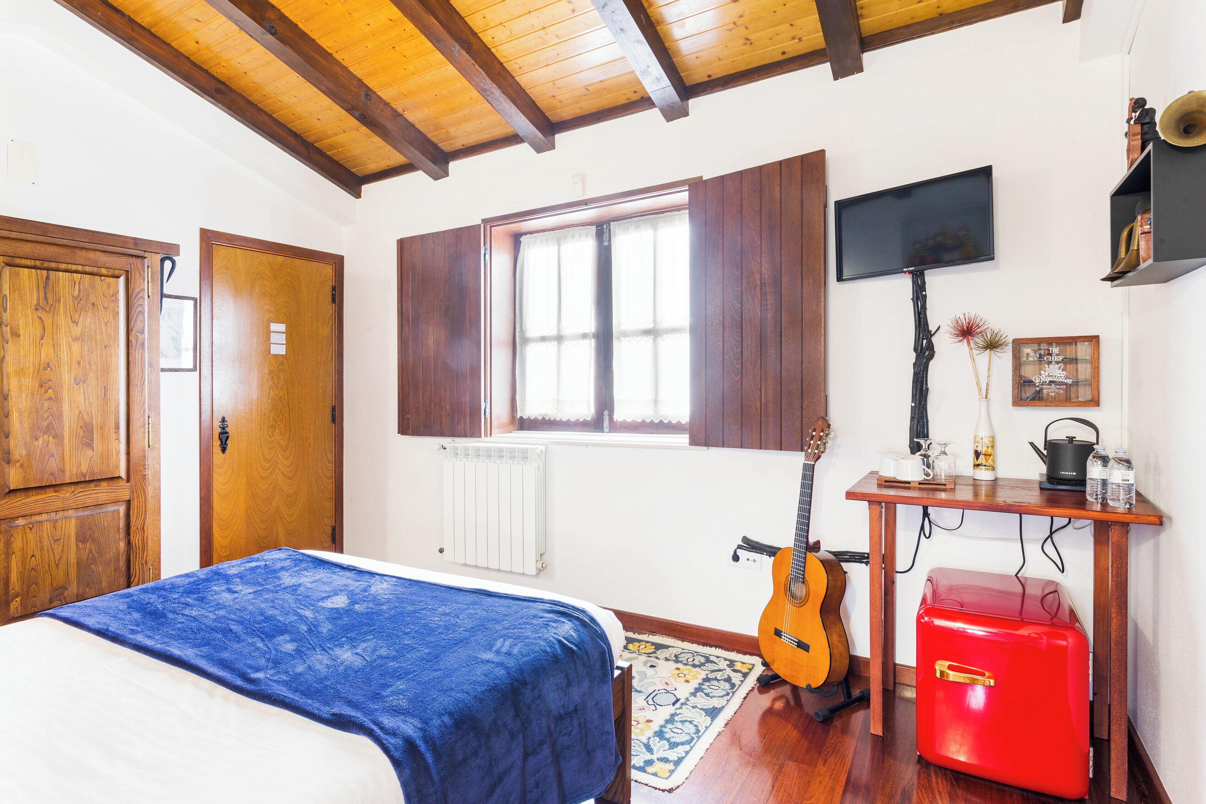 Un dormitorio con cama, una guitarra, una maleta roja, un televisor y una ventana con contraventanas de madera.