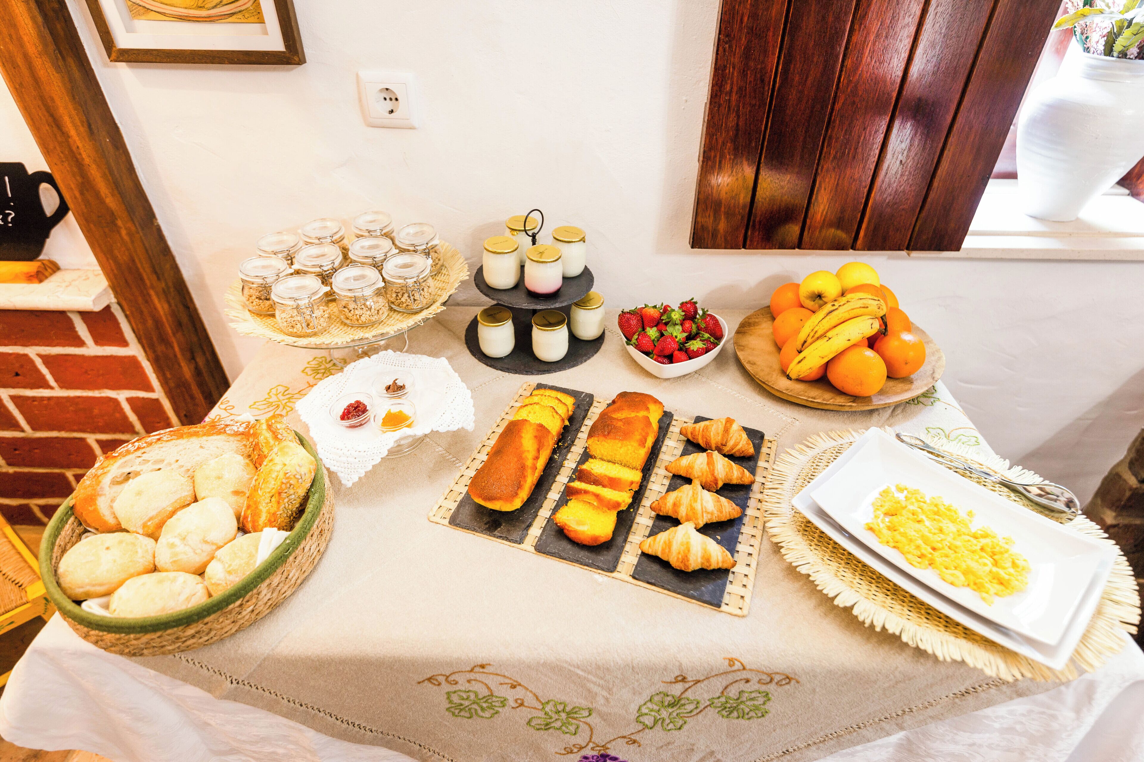 Uma mesa de buffet com diversos alimentos, incluindo pão, doces, frutas e um prato de ovos mexidos.