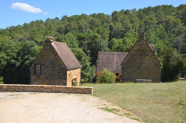 Deux bâtiments en pierre, dotés de toits à pignons, entourés d’une zone herbeuse et d’une forêt dense.