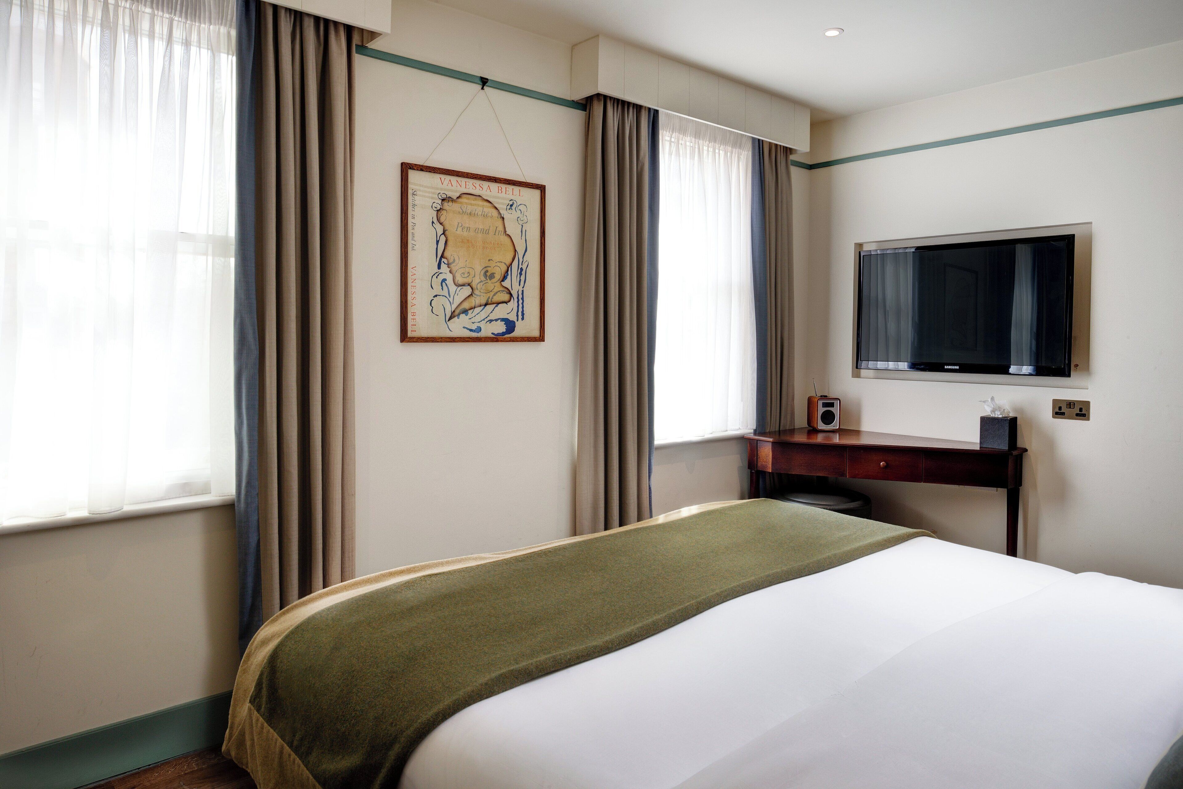 A hotel room with a bed, a desk with a flat-screen TV, a framed artwork on the wall, and a window with curtains.