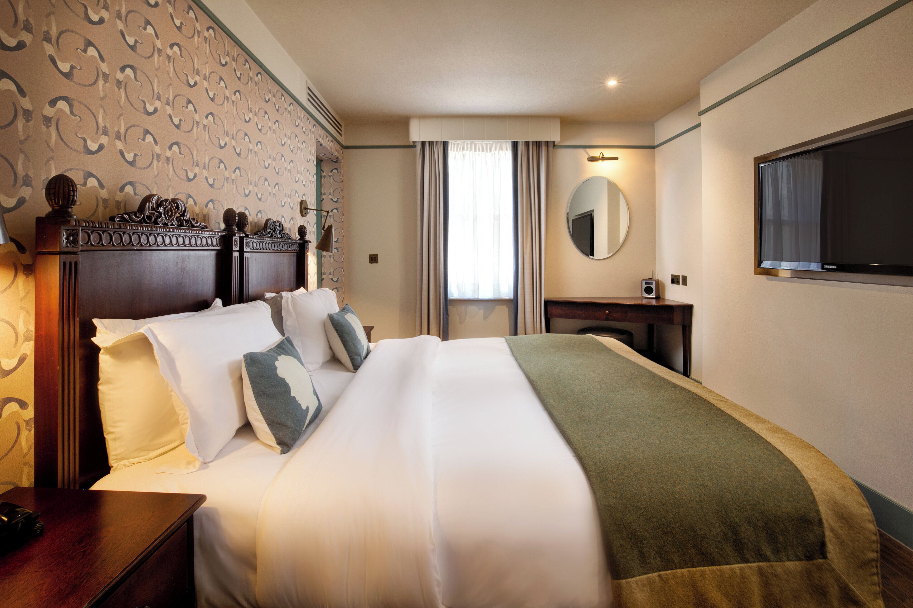 Una habitación de hotel con una cama grande, una mesita de noche con lámpara, un aparador, un espejo y un televisor en la pared.