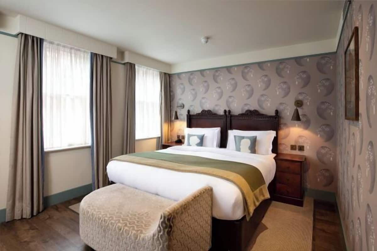 A hotel room with a large bed, a bedside table, a chair, and a patterned wall.