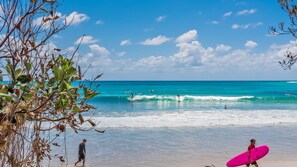 Una playa con agua azul cristalina, gente practicando surf y algunos surfistas caminando por la orilla.