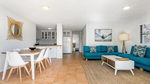 Een moderne woonkamer met een blauwgroene bank, een eethoek met witte stoelen, een keuken met witte kastjes en een strandafbeelding aan de muur.