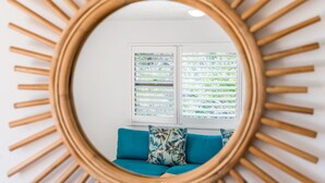 Een kamer met een ronde spiegel aan de muur, een raam met zonwering en een blauwe bank met gestreepte kussens.