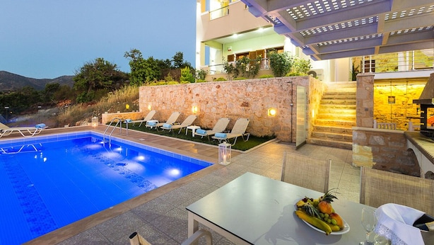 Une piscine avec des chaises longues, une table garnie de fruits et de verres, et un bâtiment doté d’une façade en pierre et d’un balcon.