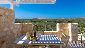Une table avec une bouteille, des verres et des fruits, placée sur un balcon doté d’un mur en pierre et offrant une vue sur un vignoble.