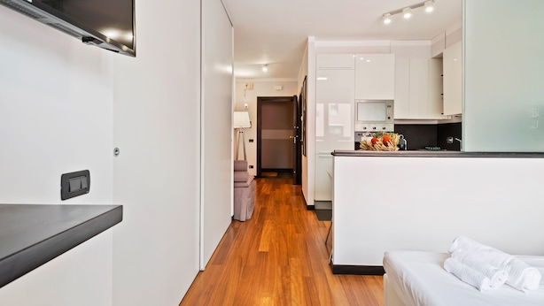 Una habitación de hotel moderna con piso de madera, una cama con ropa blanca, una kitchenette con gabinetes blancos y una pequeña zona de estar con una silla y una mesa.