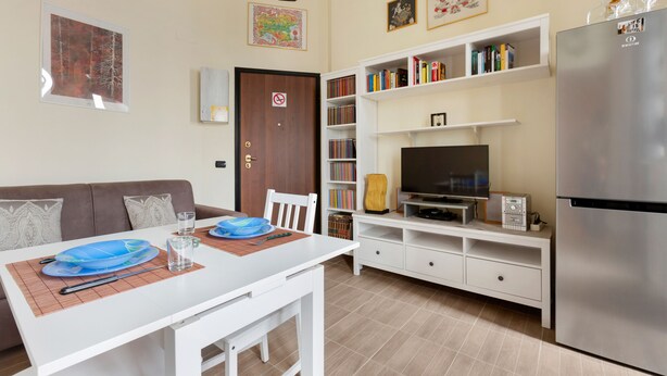 A modern kitchen with a white countertop, a built-in oven, and a refrigerator. There is a dining area with a white table and chairs, and a bookshelf filled with books. - Gambara Cosy Apartment (Milan)