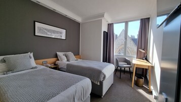 Ein Hotelzimmer mit zwei Betten, einem Schreibtisch, einem Stuhl, einem Fenster mit Vorhängen und einem gerahmten Bild an der Wand.