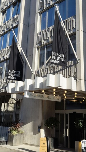 The exterior of Lancaster Hall Hotel in London, featuring a modern facade with large windows and black flags hanging from the building. - Lancaster Hall Hotel (London)