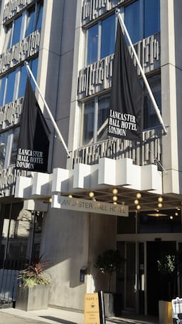 The exterior of Lancaster Hall Hotel in London, featuring a modern facade with large windows and black flags hanging from the building.