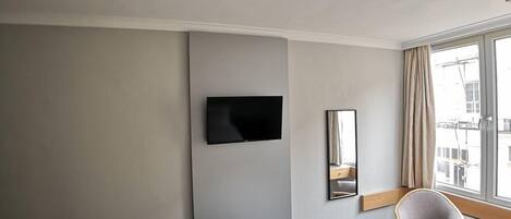 A hotel room with a flat-screen TV mounted on the wall, a mirror, a chair, and a large window with curtains.
