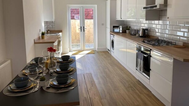 A modern kitchen with white cabinets, a built-in oven, and a dining area with a table set for four. - Beautiful Poole House 5 mins to Sandbanks +Netflix (Poole)