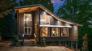 Une cabane en rondins à deux étages, dotée d’une véranda, de grandes fenêtres et d’un intérieur bien éclairé.